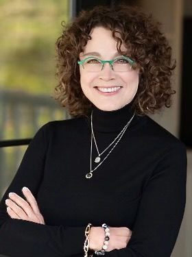 A woman with curly hair wearing glasses and smiling.